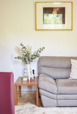 Ein Wohnzimmer mit grauem Sofa, einem kleinen Holztisch mit Pflanzenvasе und einem gerahmten Bild an der Wand.