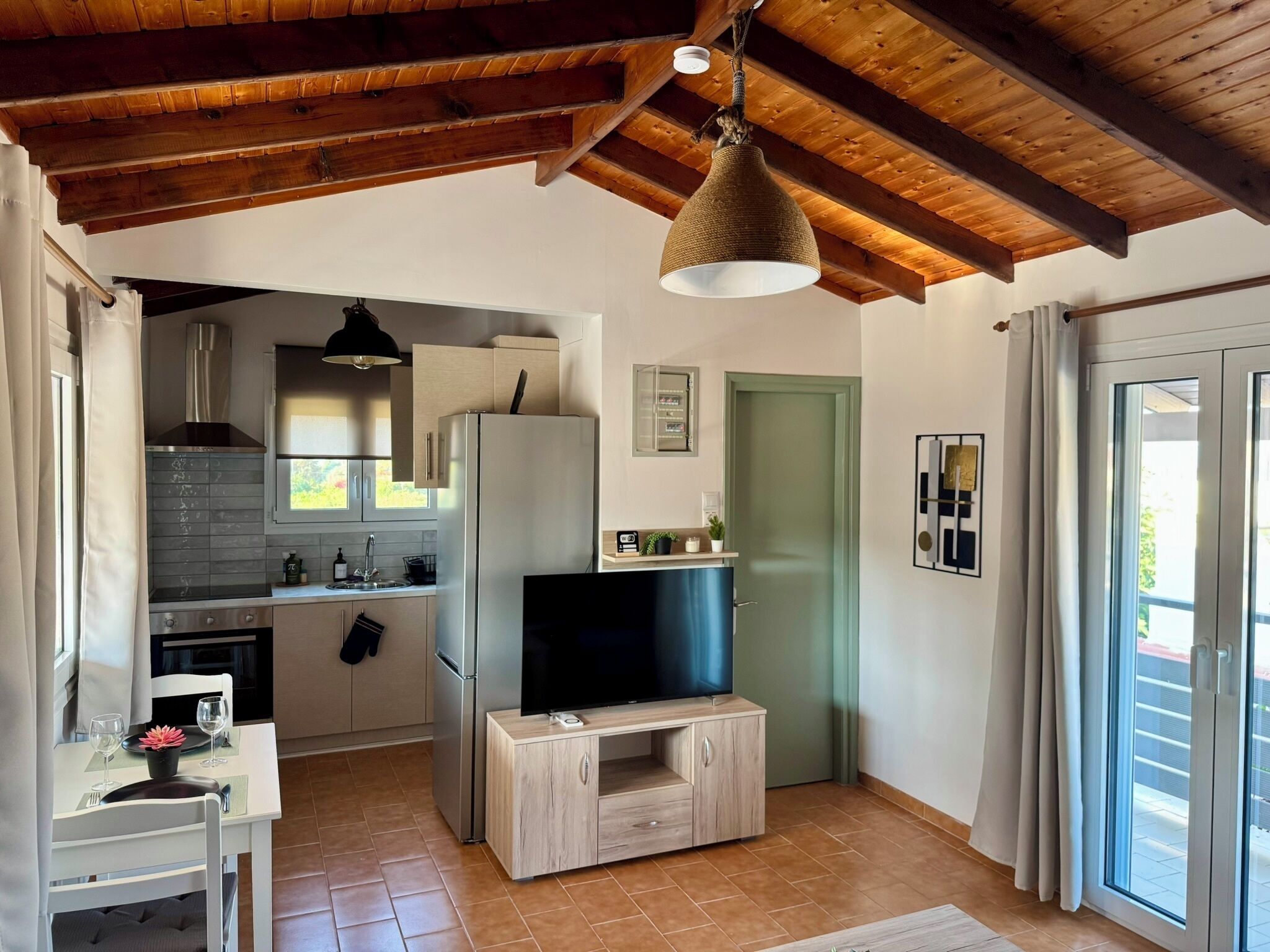 Una cocina moderna con horno integrado, un refrigerador, una mesa de comedor de madera con sillas y un televisor sobre un soporte de madera.