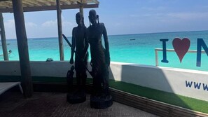A beachside view with a statue of two figures, a covered area, and a sign with 'I ♥ N' and a website address.
