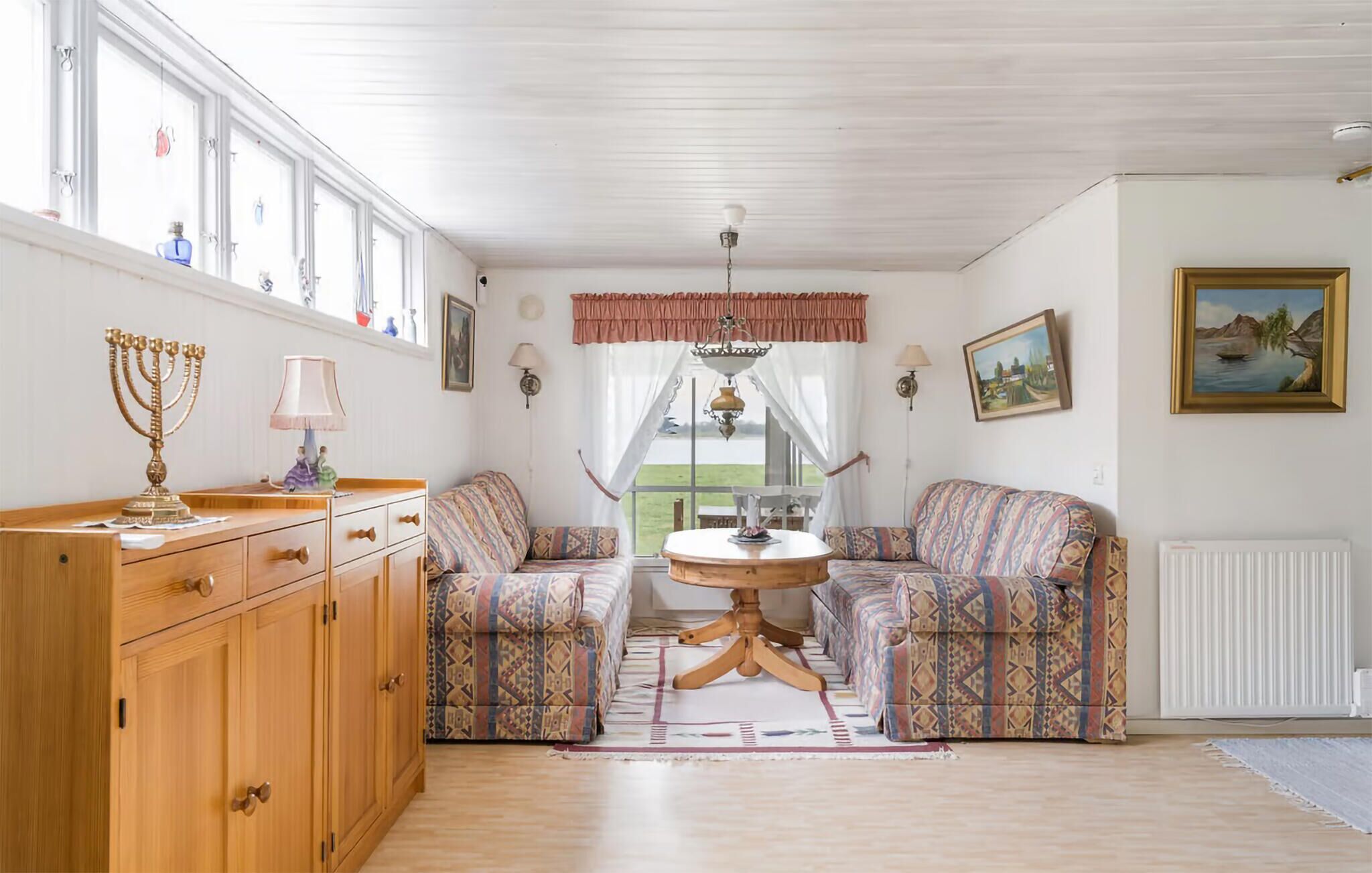 A room with a wooden cabinet, patterned sofa, a round table, and a view of the outdoors.