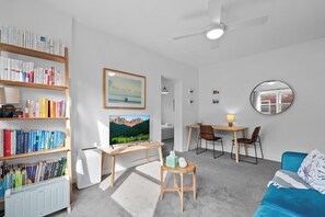 Ein modernes Wohnzimmer mit Bücherregal, Fernseher, Schreibtisch mit Stühlen, rundem Spiegel und blauem Sofa.