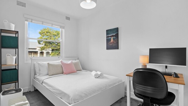 Ein Schlafzimmer mit Bett, Schreibtisch mit Computer, Stuhl, Fenster mit Blick und einem an der Wand hängenden Bild.