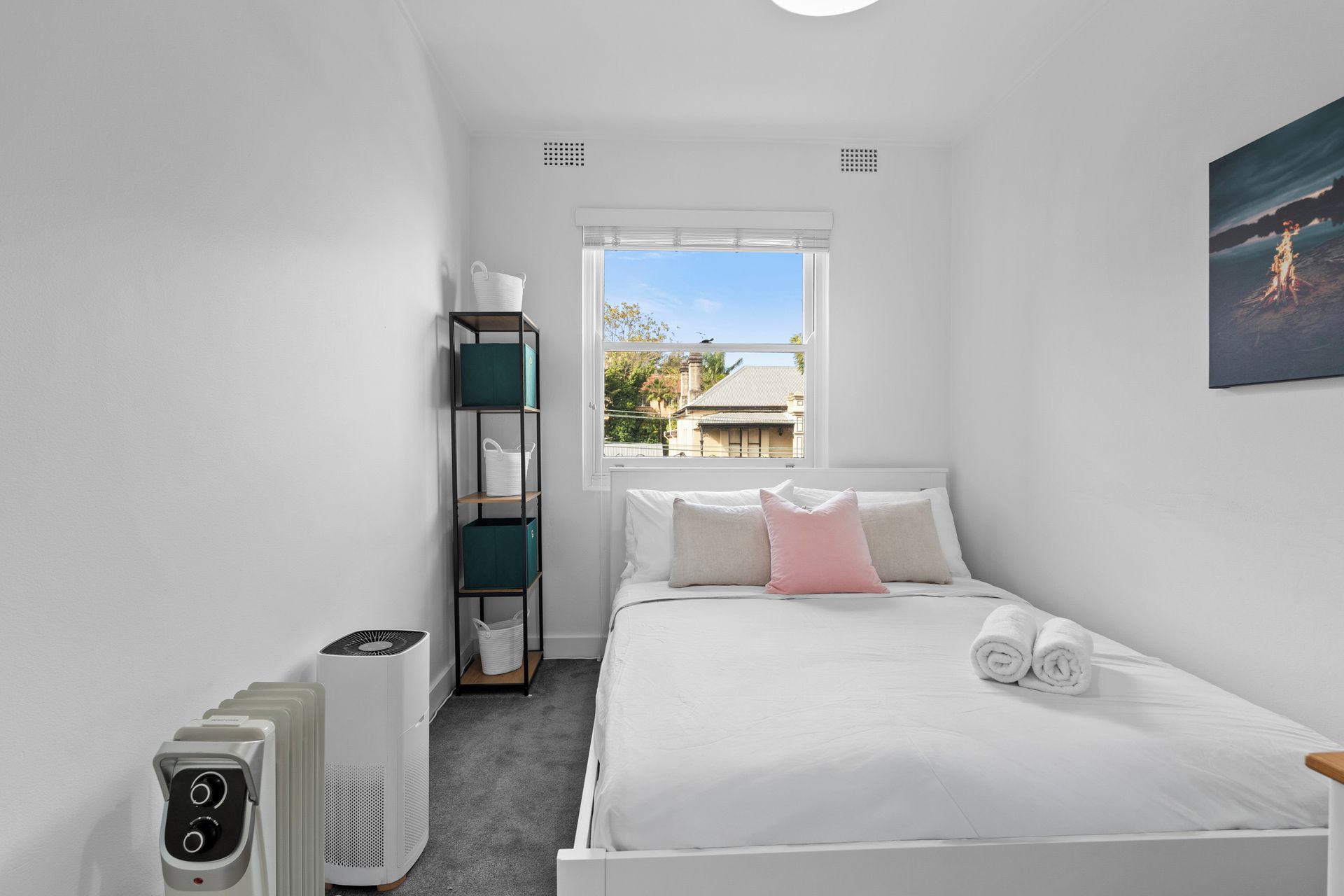 A bedroom with a bed, a window, a heater, and a shelf unit.
