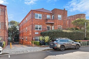 A multi-story brick building with a flat roof, a car parked in front, and a hedge in the foreground. - Bright 2BDR Bondi Junction Home with Parking (Bondi Junction)