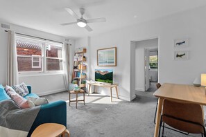 A modern living room with a blue sofa, a wooden coffee table, a bookshelf, a painting, and a dining area with a table and chairs. - Bright 2BDR Bondi Junction Home with Parking (Bondi Junction)