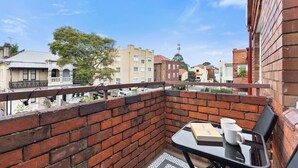 Ein Balkon mit Ziegelmauer, einem Tisch mit einem Buch und zwei Tassen sowie Blick auf umliegende Gebäude.