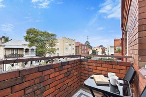 Uma varanda com um muro de tijolos, uma mesa com um livro e duas canecas, e vista para edifícios ao redor.