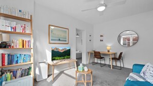 Ein modernes Wohnzimmer mit Bücherregal, Fernseher, Schreibtisch mit Stühlen, rundem Spiegel und blauem Sofa.