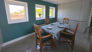 Un coin repas avec une table dressée pour quatre personnes, deux fenêtres à cadres blancs, un radiateur et une armoire blanche.