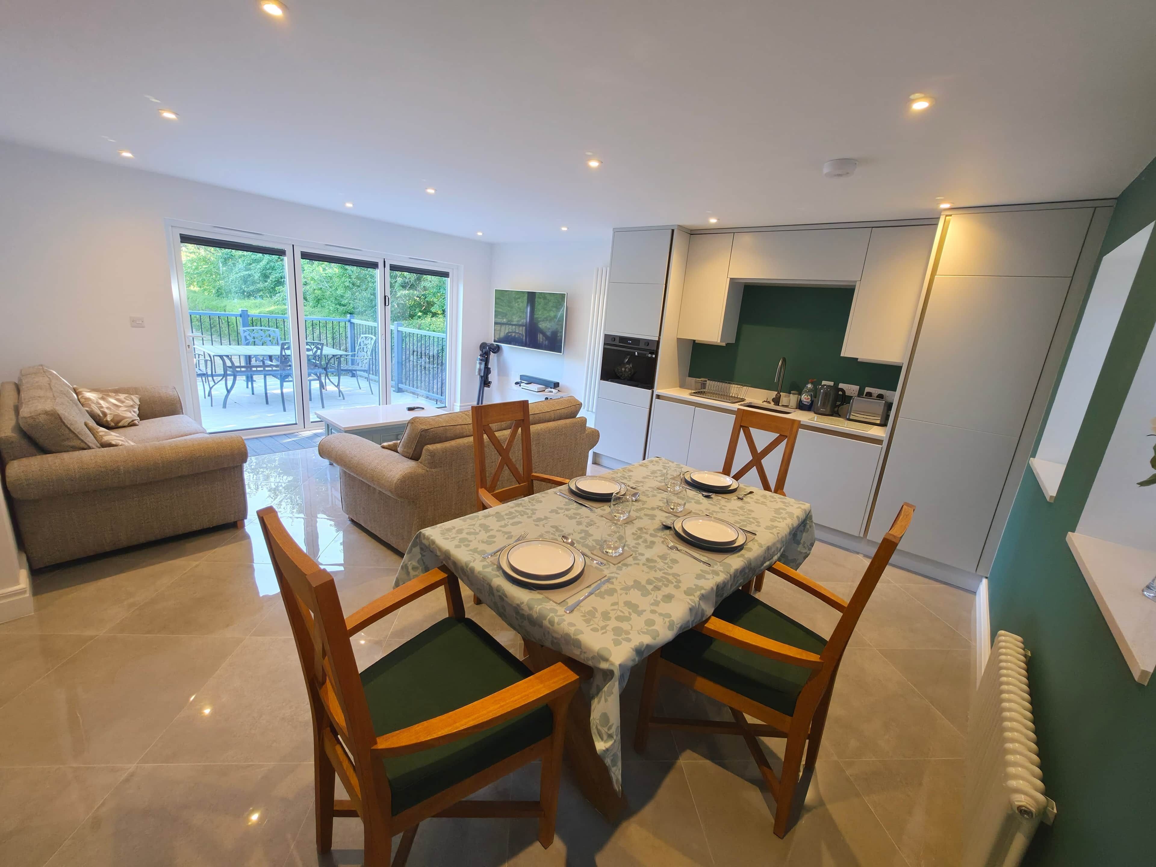 A modern living room with a dining area, a sofa, a table set for four, and a kitchen in the background.