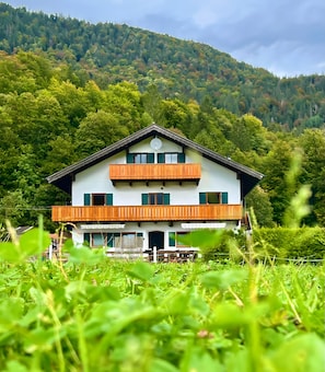 Eine zweistöckige Chalet mit Balkon, umgeben von üppiger Vegetation und einem bewaldeten Hügelhang.