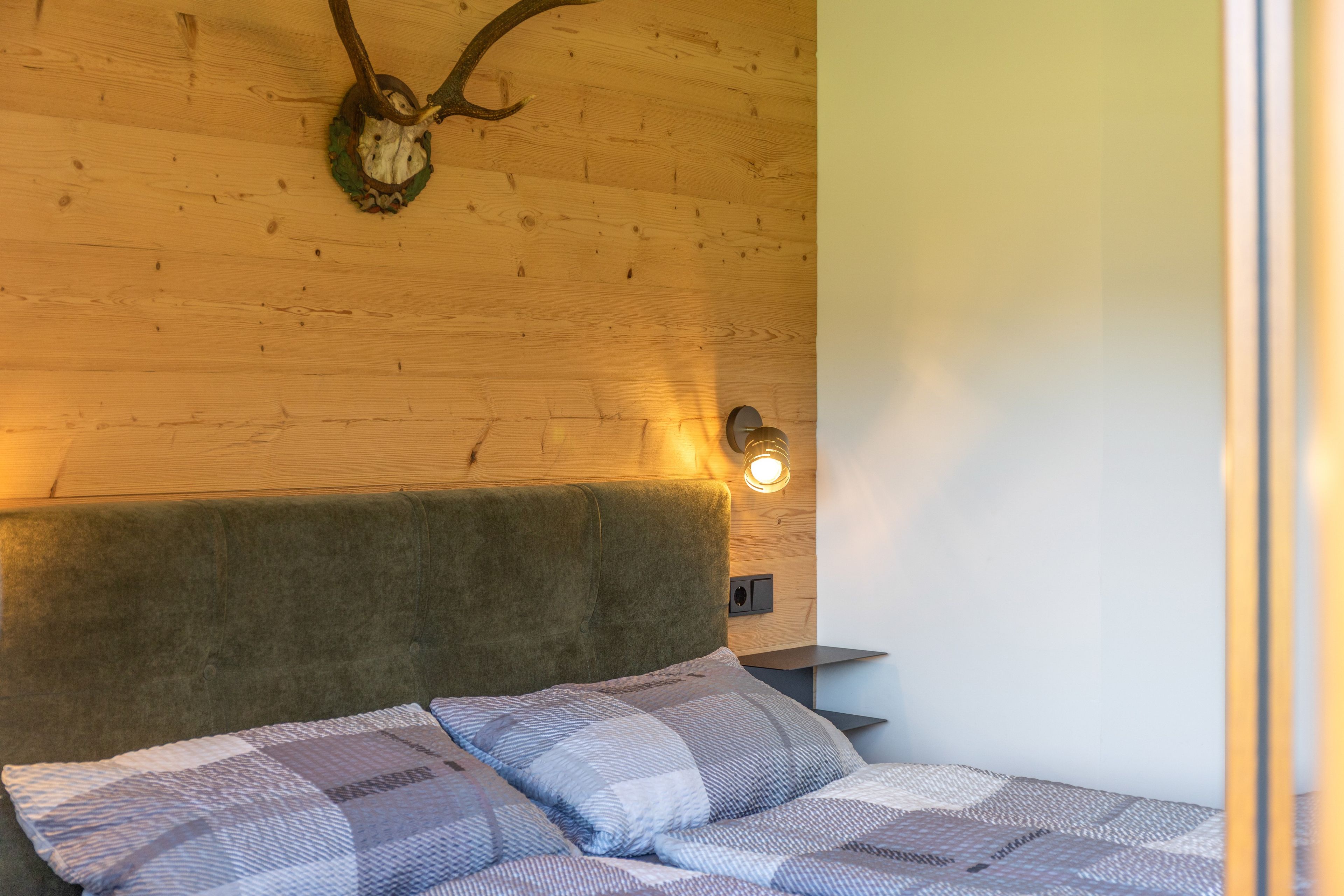 A room with a wooden headboard, a bed with patterned bedding, a wall-mounted light, and a wooden wall with a mounted animal skull.