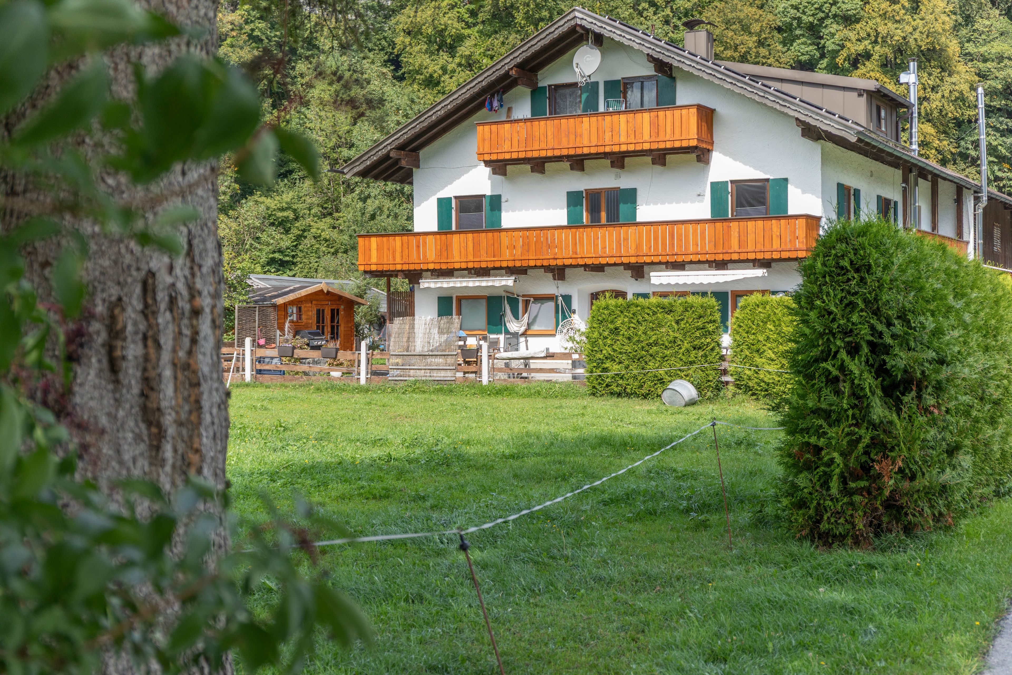 Ein zweistöckiges Gebäude mit Balkon, umgeben von viel Grün und einem eingezäunten Garten.