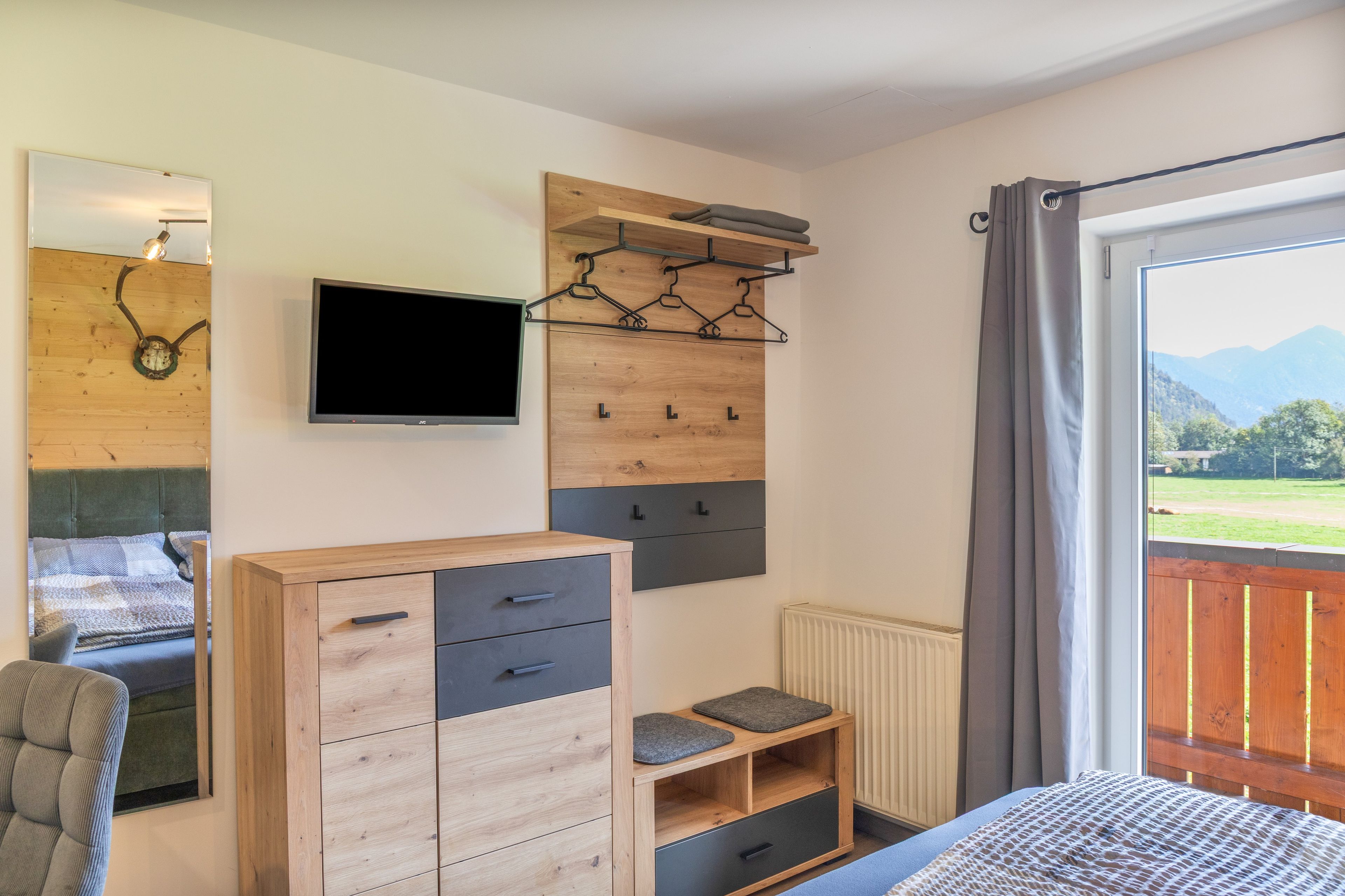 Ein Schlafzimmer mit einem Bett, einem Fernseher, einer Holzkommode, einem Hakenregal und Blick ins Freie.
