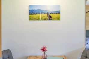 Ein gerahmtes Foto zeigt ein Feld mit Bergen im Hintergrund, einen Tisch mit Blumen in einer Vase und zwei Stühle.