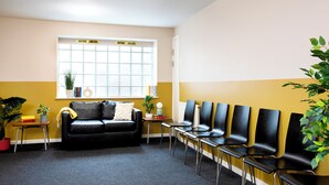A waiting area with a black leather sofa, a window with blinds, a row of black chairs, and potted plants. - Student Only Cross Court House (London)