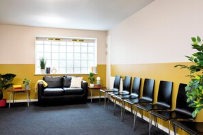 Ein Wartebereich mit einer schwarzen Ledersofa, einem Fenster mit Jalousien, einer Reihe schwarzer Stühle und Blumentöpfen.