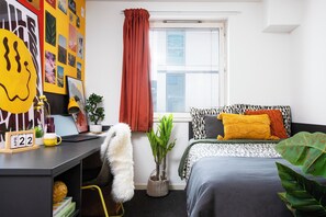 Ein modernes Schlafzimmer mit Schreibtisch, einem Bett mit gemusterten Betttextilien, einem Fenster mit Jalousien und einer Wand, die mit verschiedenen Bildern geschmückt ist.