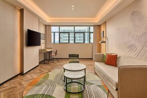A modern hotel room with a flat-screen TV, a sofa, a small round table, and a view of a building through the window.