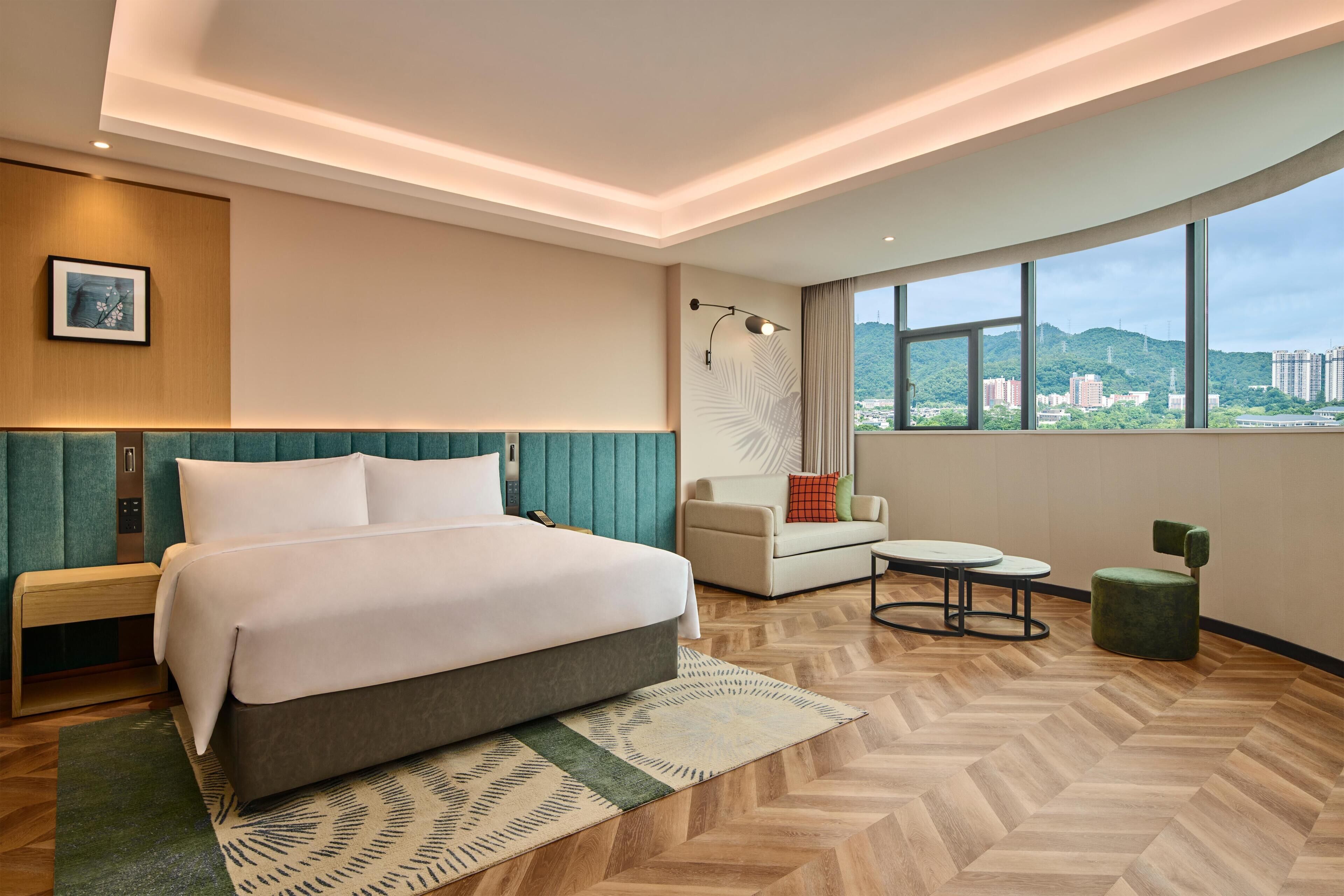 A modern hotel room with a large bed, a sofa, a small table, and a view of the city.