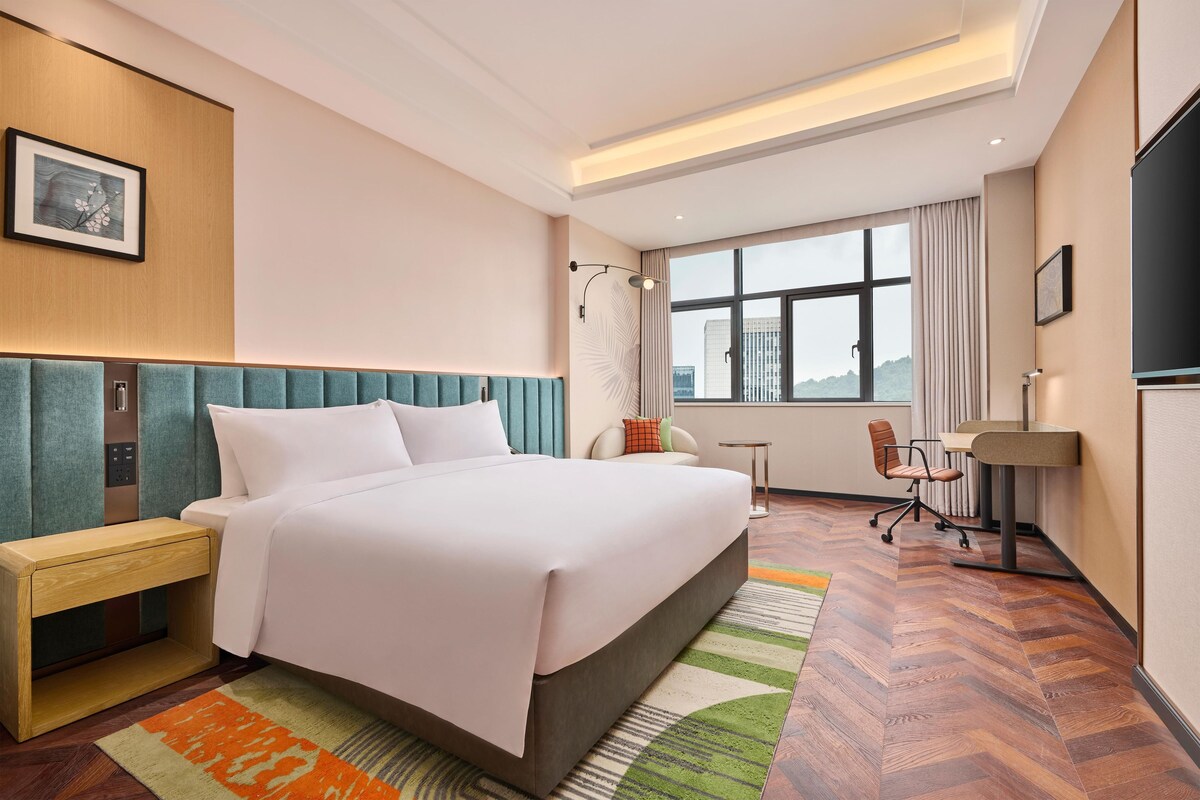 A hotel room with a bed, a desk, a chair, a TV, and a window with a city view.
