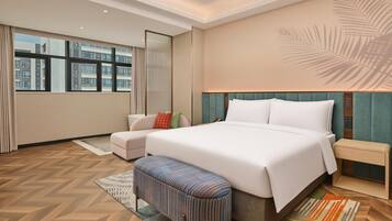 A hotel room with a large bed, a nightstand, a bench, and a view of the city through large windows.