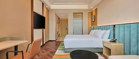 A hotel room with a bed, a desk, a chair, a small table, and a television.