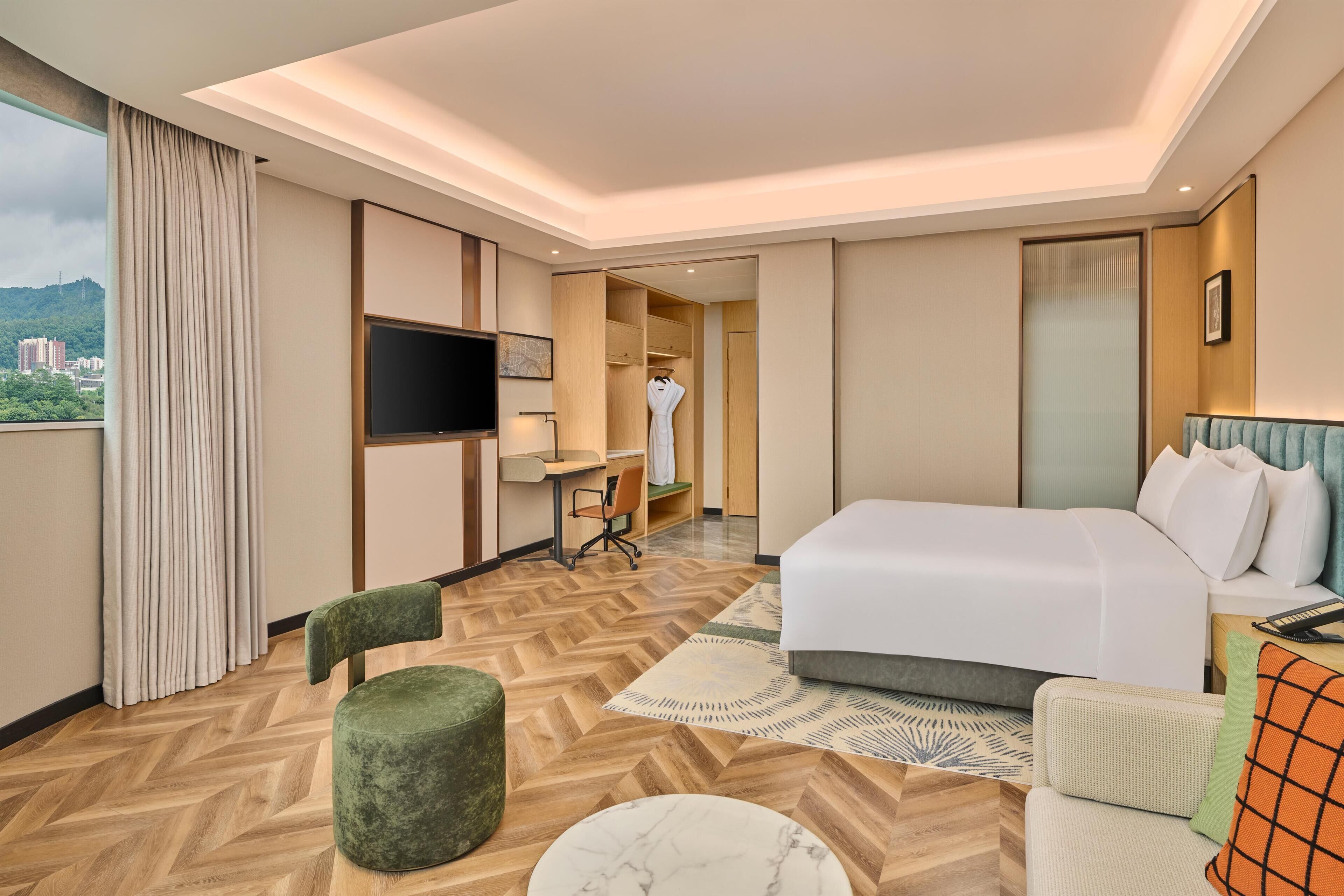 A hotel room with a bed, a desk, a chair, a television, and a view of the city.
