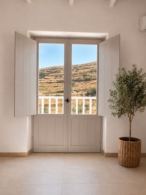A room with a door leading to a balcony with a view of a hilly landscape. - Dryope Luxury Living (Kithnos)