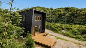 A modern black building with a wooden deck and glass doors, surrounded by lush greenery. - Log Cabin with Ocean Views | Bonfire in a Private Satoyama Forest (富津市)