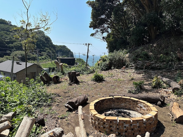 A rural outdoor setting with a brick fire pit, wooden logs, and a house in the background.