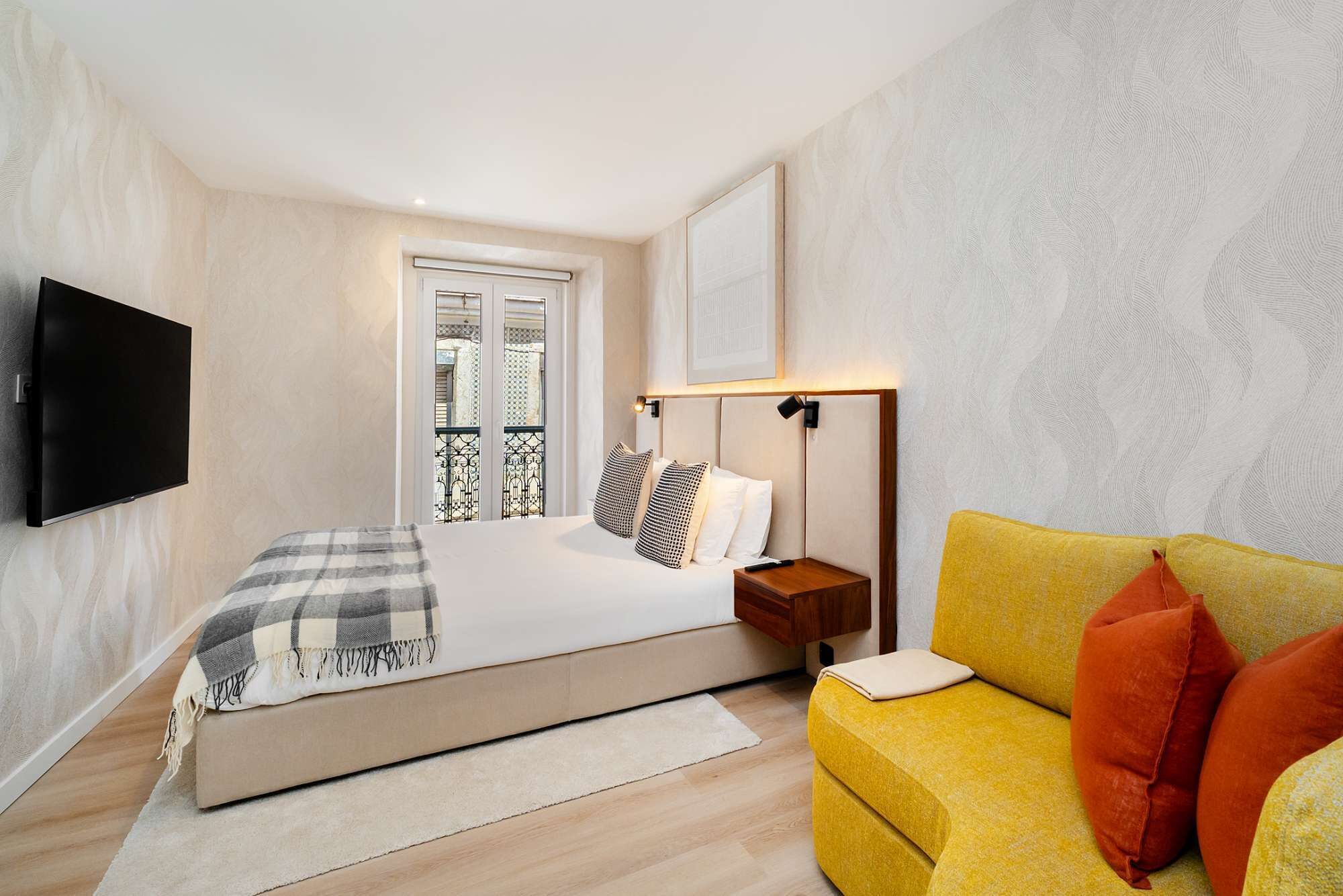A hotel room with a bed, a television, a sofa, and a window with a balcony.