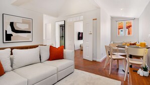 En moderne stue med en sofa, et spiseområde med stole, et bord dækket til morgenmad, og et køkken, der kan ses gennem en åben dør.