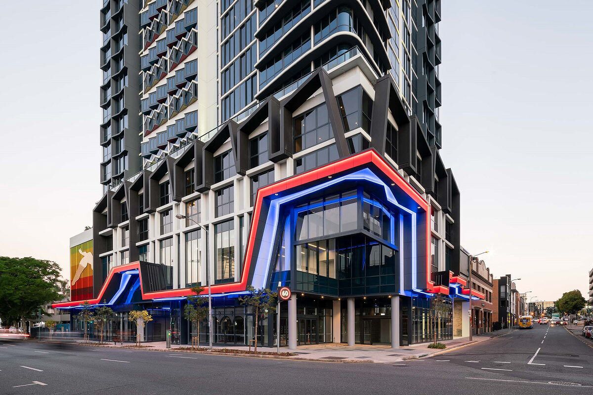 A modern building with a curved glass facade, a prominent red and blue entrance, and a large vertical banner on the left side.