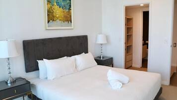 Una cama bien hecha con sábanas blancas, un cabecero oscuro, una lámpara de noche, una mesita de noche y un cuadro en la pared.