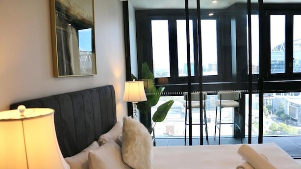 Ein Hotelzimmer mit Bett, Kissen, einer Lampe und einem großen Fenster mit Blick auf Gebäude.