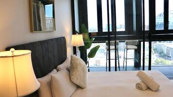 Habitación de hotel con cama, almohadas, lámpara y un ventanal con vistas a edificios.