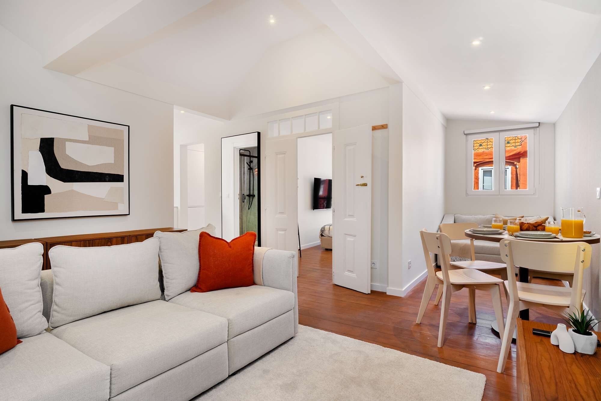 A modern living room with a sofa, a dining area with chairs, a table set for breakfast, and a kitchen visible through an open door.