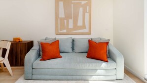 En stue med en lyseblå sofa, to orange puder, et træsidebord med en glaskrukke, og et indrammet abstrakt kunstværk på væggen.