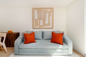Ein Wohnzimmer mit einem hellblauen Sofa, zwei orangenen Kissen, einem Holznachttisch mit einem Glasbehälter und einem gerahmten abstrakten Kunstwerk an der Wand.
