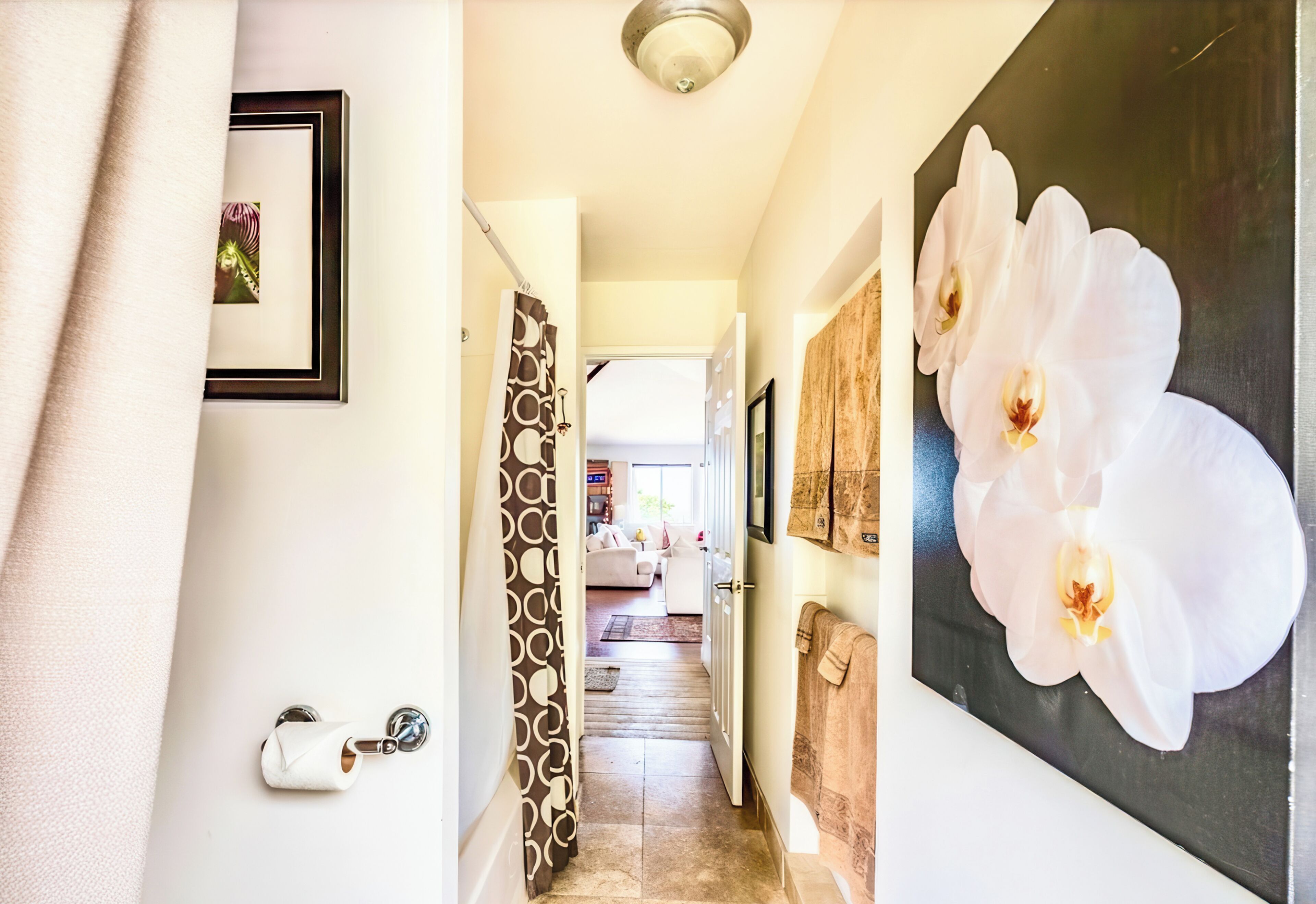 Un pasillo con dispensador de papel higiénico, cortina de ducha estampada y un cuadro grande de orquídeas.