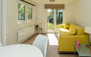 Ein heller Raum mit einem gelben Sofa, einem weißen Tisch und einem Fenster mit Blick auf Grünflächen.