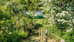 Un chemin mène à un bâtiment à la façade verte et blanche, entouré d’une végétation luxuriante et d’arbustes à fleurs blanches.