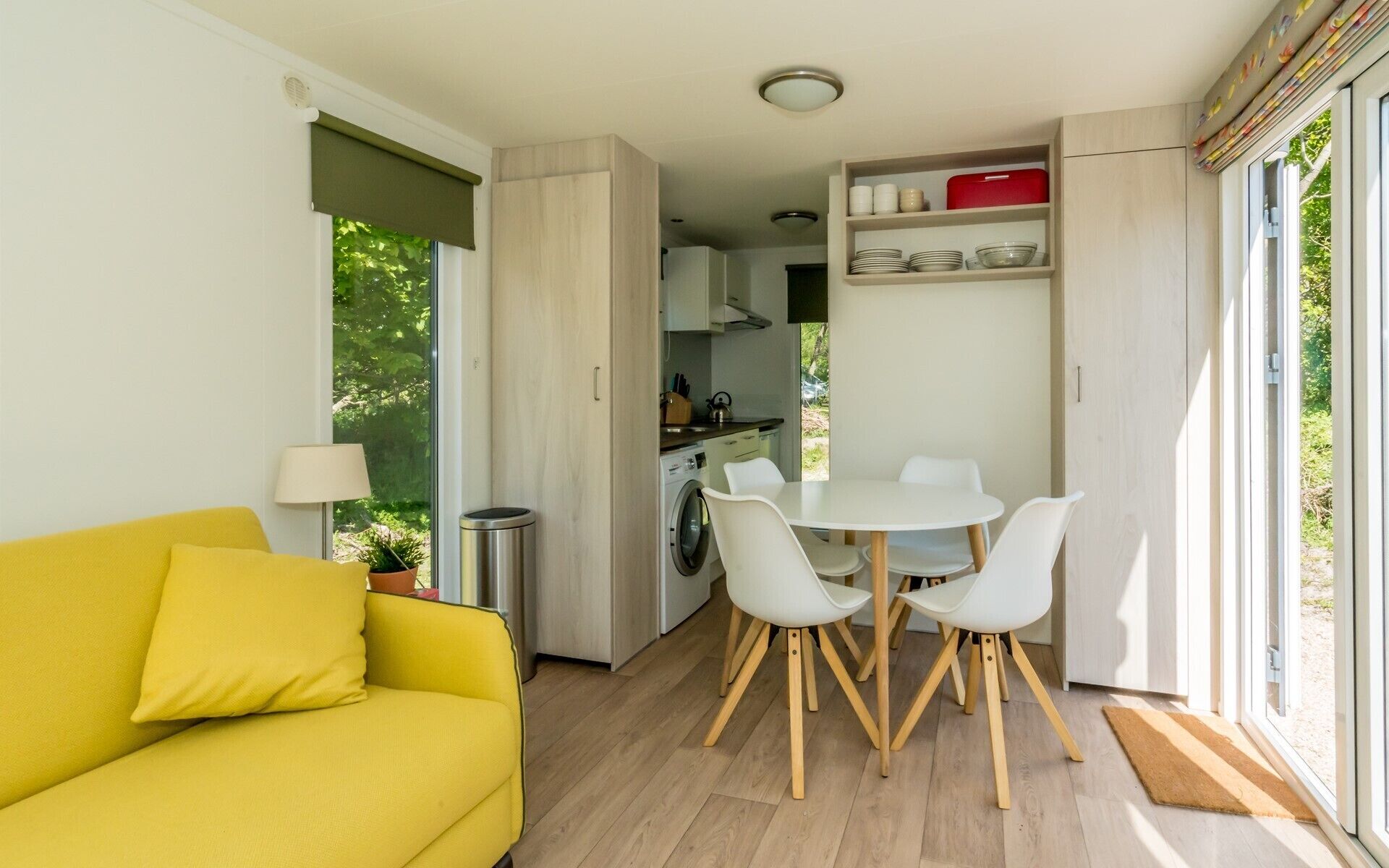 A compact living space with a yellow sofa, a round dining table with white chairs, a kitchen area, and a view of greenery through the windows.