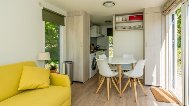 Un espace de vie compact comprenant un canapé jaune, une table de salle à manger ronde avec des chaises blanches, un coin cuisine et une vue sur la verdure par les fenêtres.