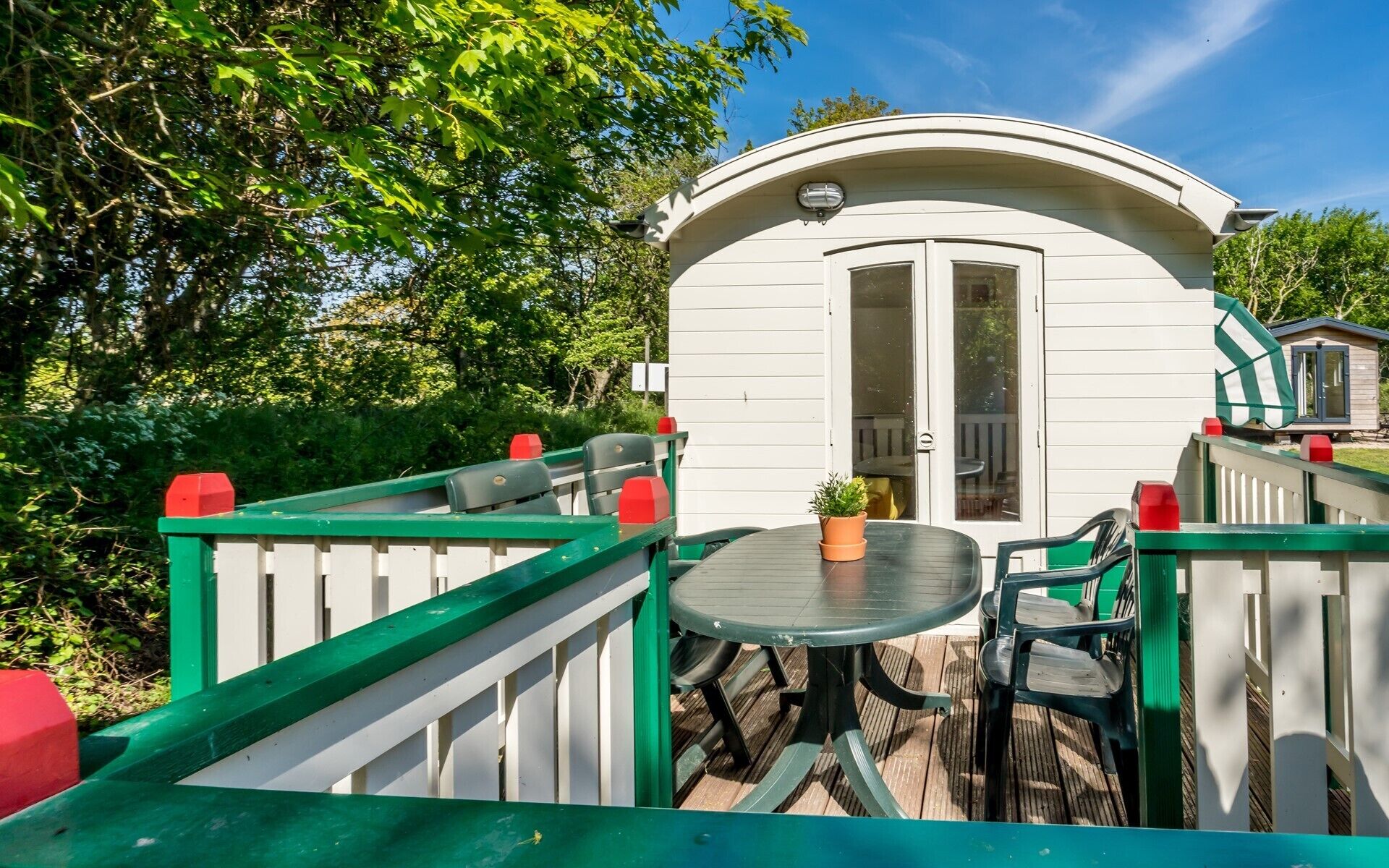A white mobile home with a curved roof, a small deck with a table and chairs, surrounded by greenery.