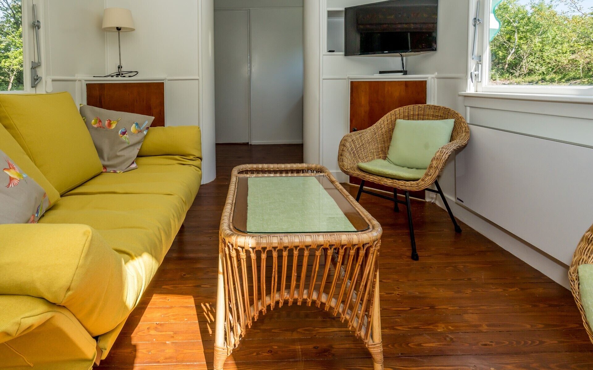 A room with a sofa, a wicker coffee table, a wicker armchair, a television mounted on the wall, and a window with a view of trees.