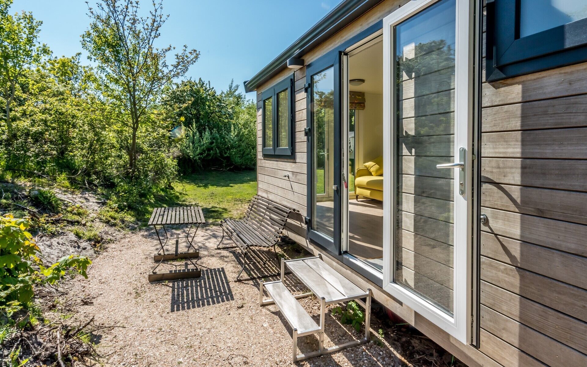 Ein modernes Haus mit Terrasse, Sitzgelegenheiten im Freien und einem sichtbaren Innenraum mit einem gelben Sessel.