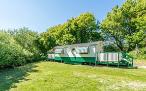Ein Mobilheim mit grüner Außenwand und Streifenmarkisen, umgeben von üppigem Grün und Bäumen.