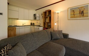 Une cuisine moderne avec des armoires blanches, un four à micro-ondes et une machine à café. On y trouve une étagère en bois ornée d’objets décoratifs et un lampadaire. Un tableau représentant une scène de plage est accroché au mur.